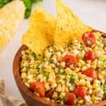 A wooden bowl filled with corn salsa, cherry tomatoes, herbs, and two tortilla chips, with corn on the cob and greens in the background.