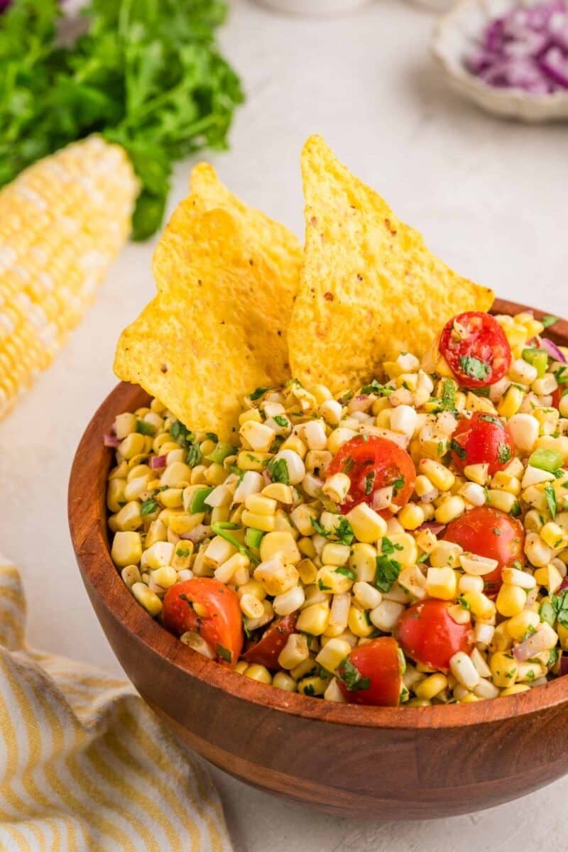 A wooden bowl filled with corn salsa, cherry tomatoes, herbs, and two tortilla chips, with corn on the cob and greens in the background.