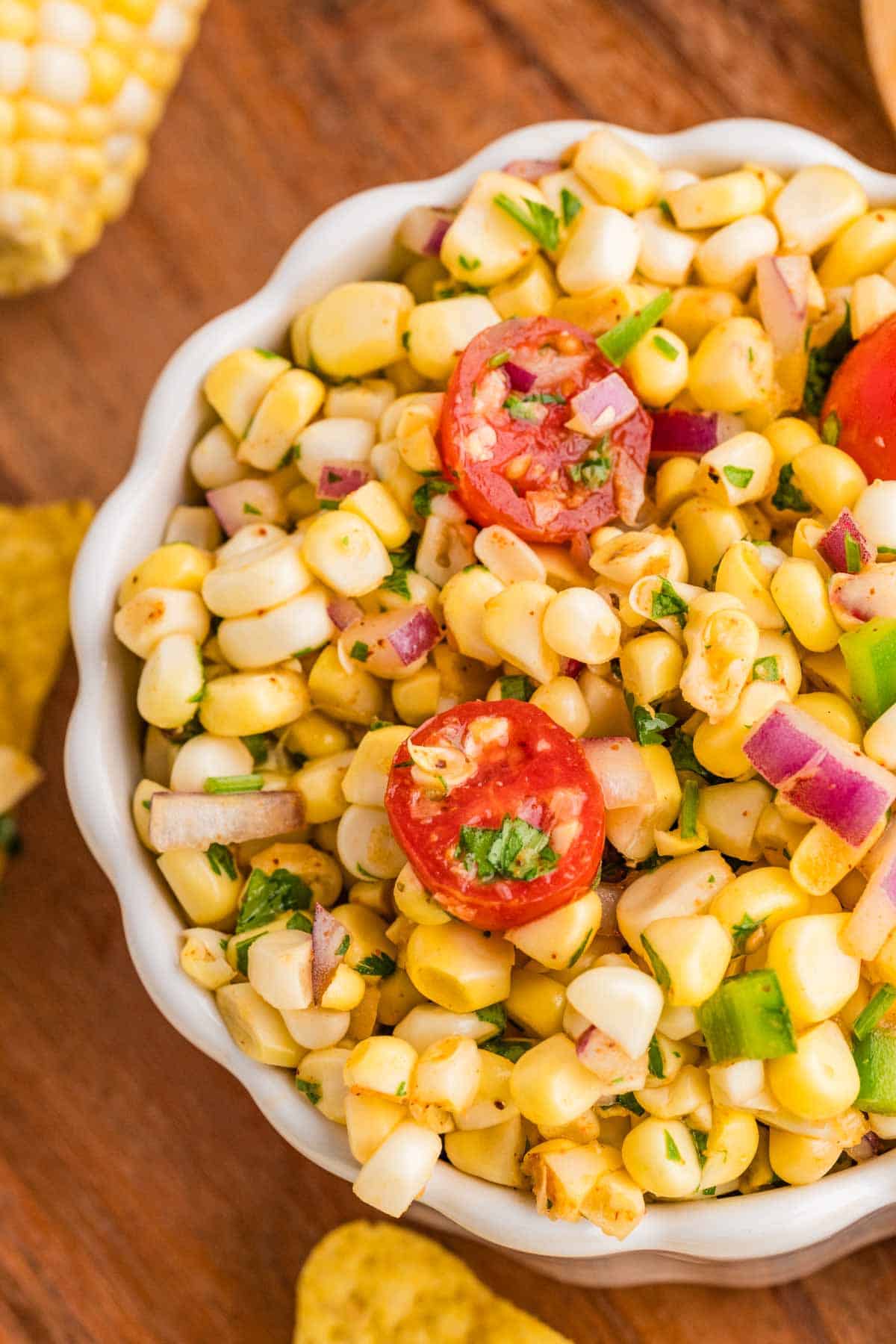 A bowl of corn salad with diced red onion, chopped green pepper, cilantro, and halved cherry tomatoes on a wooden surface.