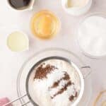 Baking ingredients on a countertop including flour, cocoa, and sugar in bowls, eggs, coffee, oil, cream, and a whisk, with a sifter over a glass bowl.