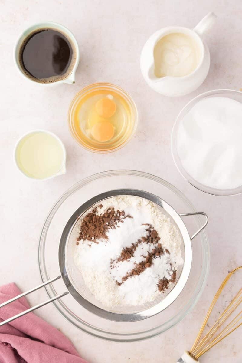 Baking ingredients on a countertop including flour, cocoa, and sugar in bowls, eggs, coffee, oil, cream, and a whisk, with a sifter over a glass bowl.