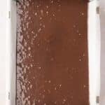 A rectangular baking pan lined with parchment paper filled with unbaked chocolate brownie batter.