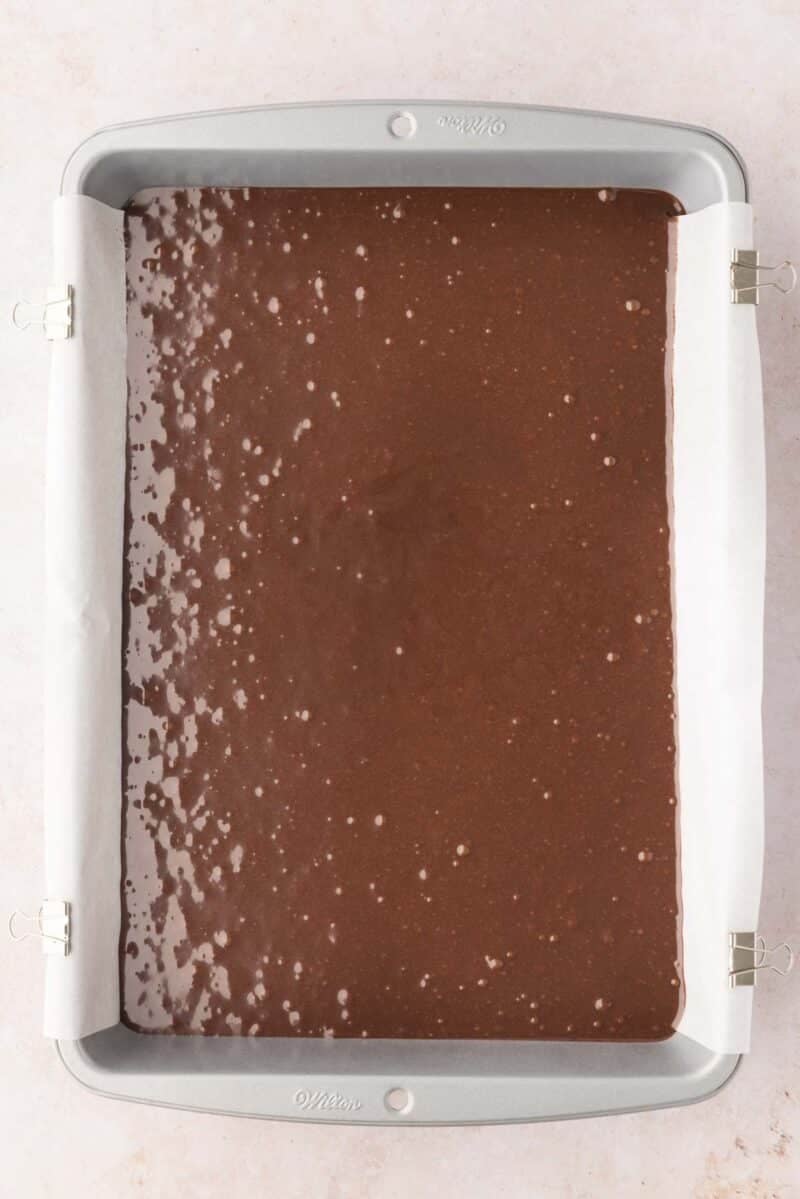 A rectangular baking pan lined with parchment paper filled with unbaked chocolate brownie batter.