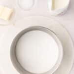 A saucepan filled with granulated sugar is on a marble surface, surrounded by small bowls of butter, vanilla, salt, and a jug of cream.