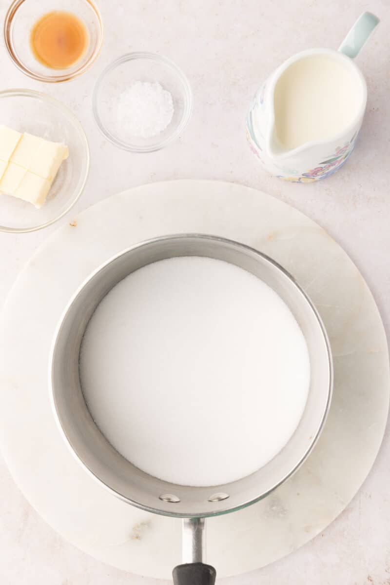 A saucepan filled with granulated sugar is on a marble surface, surrounded by small bowls of butter, vanilla, salt, and a jug of cream.