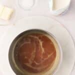 A saucepan with caramelized sugar sits on a marble trivet, surrounded by small bowls of vanilla, salt, butter, and a jug of cream.