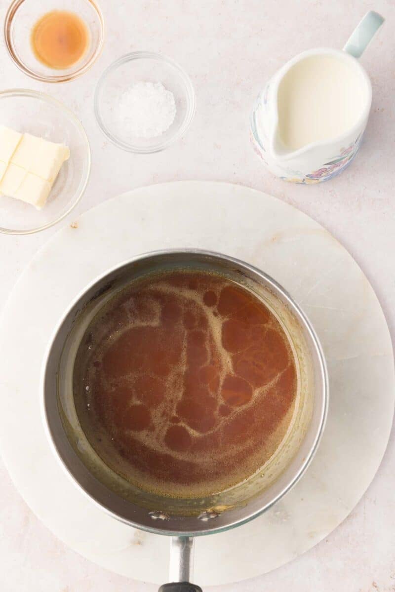 A saucepan with caramelized sugar sits on a marble trivet, surrounded by small bowls of vanilla, salt, butter, and a jug of cream.