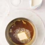 A saucepan with melted brown sugar and butter sits on a marble surface. Nearby are a small pitcher of cream, a bowl of salt, and a dish with liquid, likely vanilla extract.