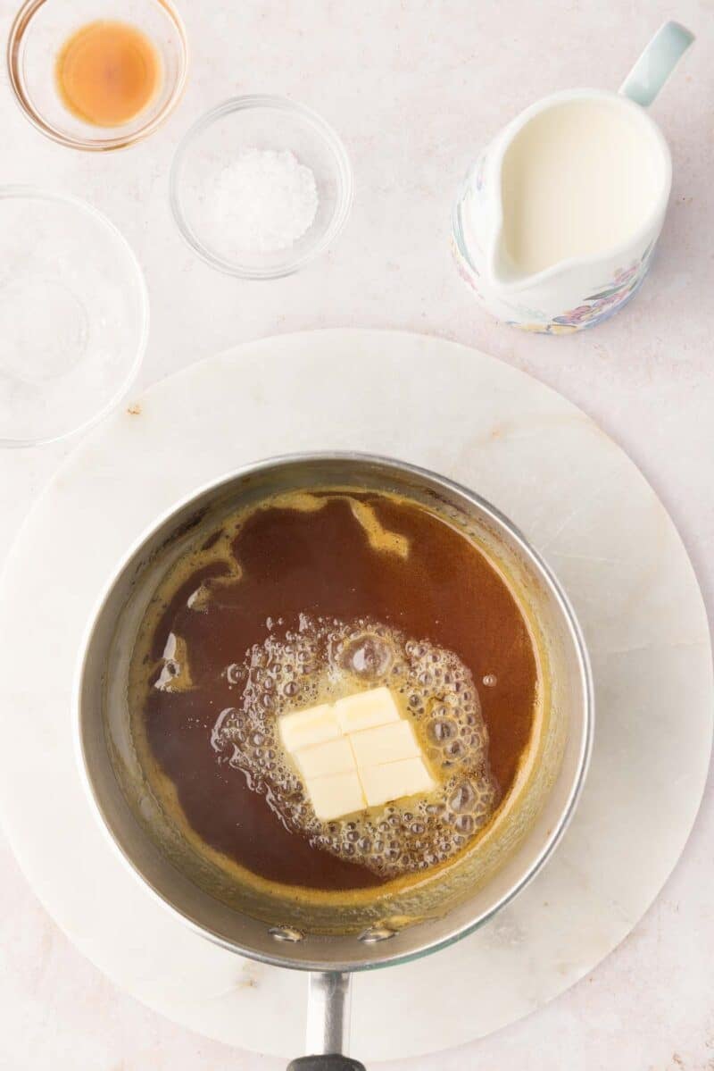 A saucepan with melted brown sugar and butter sits on a marble surface. Nearby are a small pitcher of cream, a bowl of salt, and a dish with liquid, likely vanilla extract.