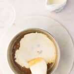 A saucepan contains a creamy mixture being stirred with a spatula; a pitcher of cream and a small bowl of vanilla extract are nearby on a light countertop.