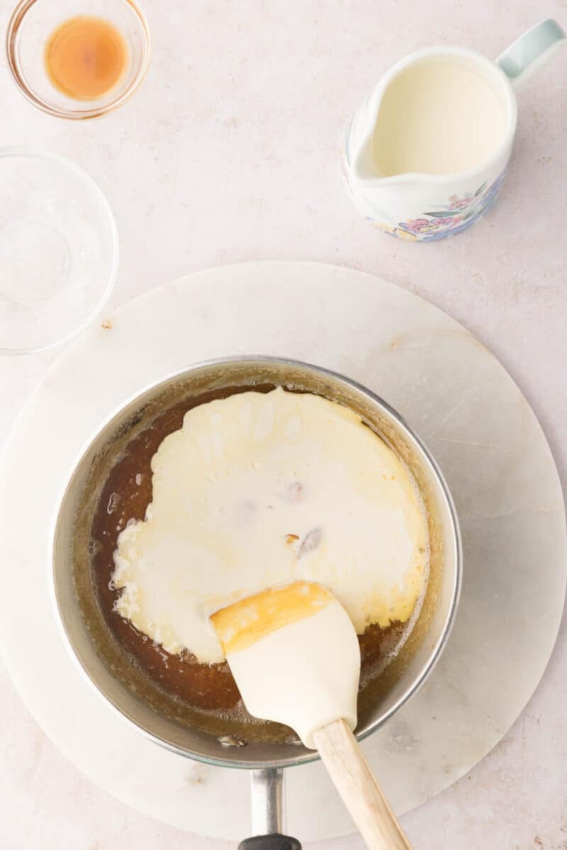 A saucepan contains a creamy mixture being stirred with a spatula; a pitcher of cream and a small bowl of vanilla extract are nearby on a light countertop.