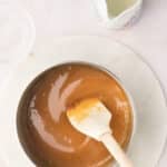 A saucepan filled with caramel sauce being stirred with a spatula, surrounded by empty glass bowls and a small pitcher on a light surface.