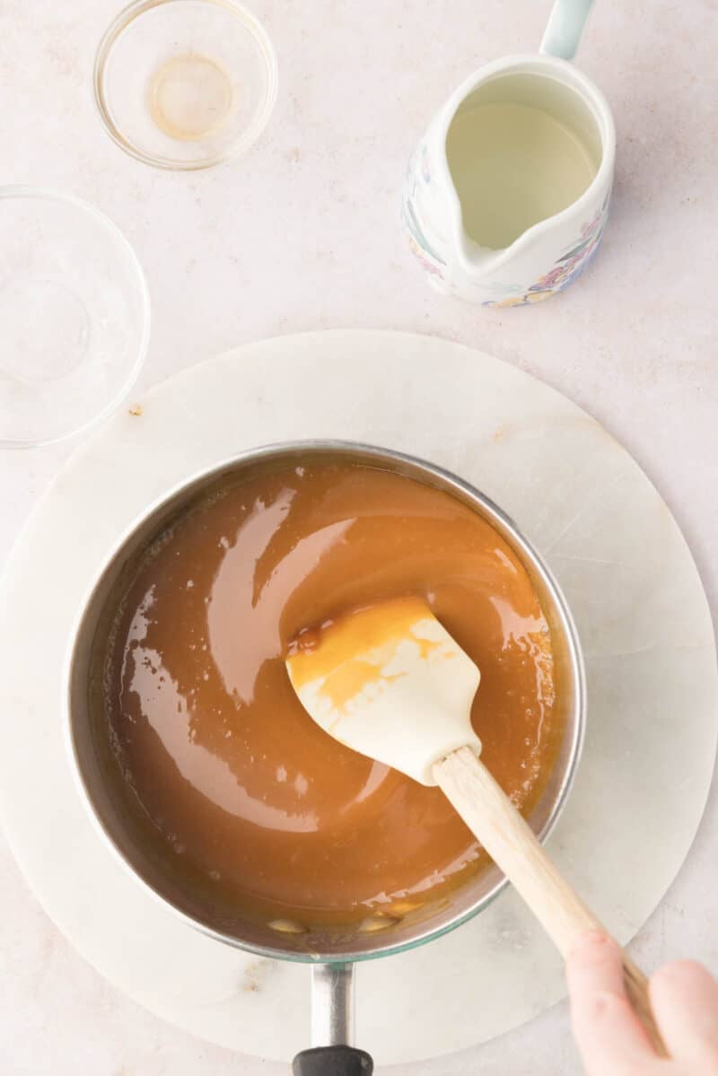 A saucepan filled with caramel sauce being stirred with a spatula, surrounded by empty glass bowls and a small pitcher on a light surface.