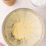 A mixing bowl with creamed butter, a jar of caramel sauce, and a bowl of powdered sugar on a countertop.
