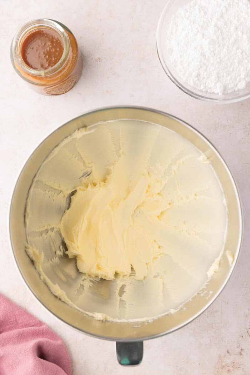 A mixing bowl with creamed butter, a jar of caramel sauce, and a bowl of powdered sugar on a countertop.