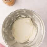 A mixing bowl with creamy white frosting and a small jar of caramel sauce on a light countertop.