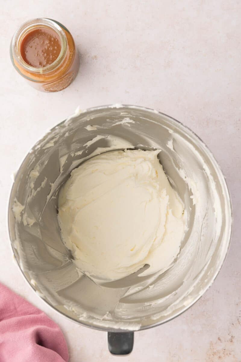 A mixing bowl with creamy white frosting and a small jar of caramel sauce on a light countertop.