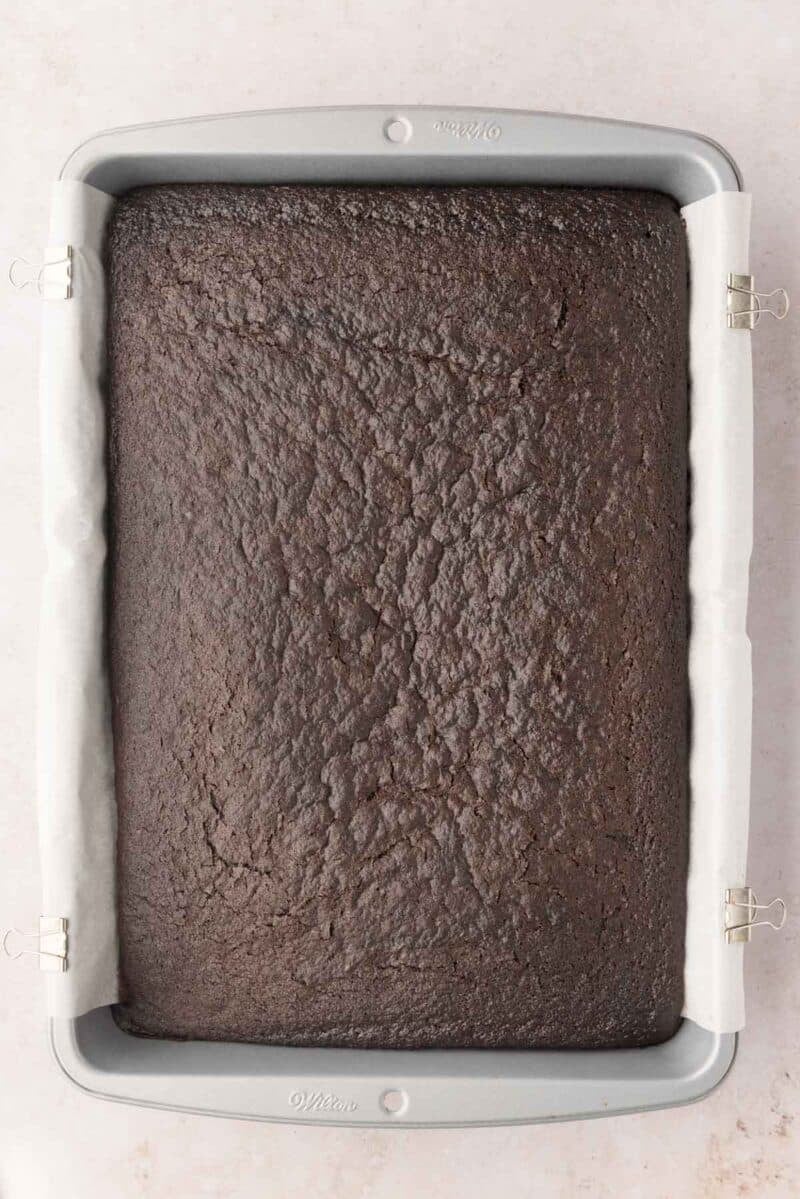 A rectangular pan lined with parchment paper holds a freshly baked, uncut chocolate cake.