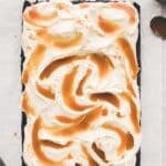 A rectangular cake topped with swirled white frosting and caramel sauce, placed on parchment paper with a spatula and a jar of caramel nearby.