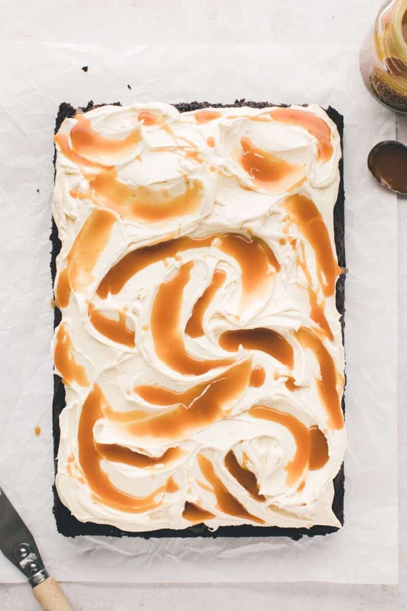 A rectangular cake topped with swirled white frosting and caramel sauce, placed on parchment paper with a spatula and a jar of caramel nearby.
