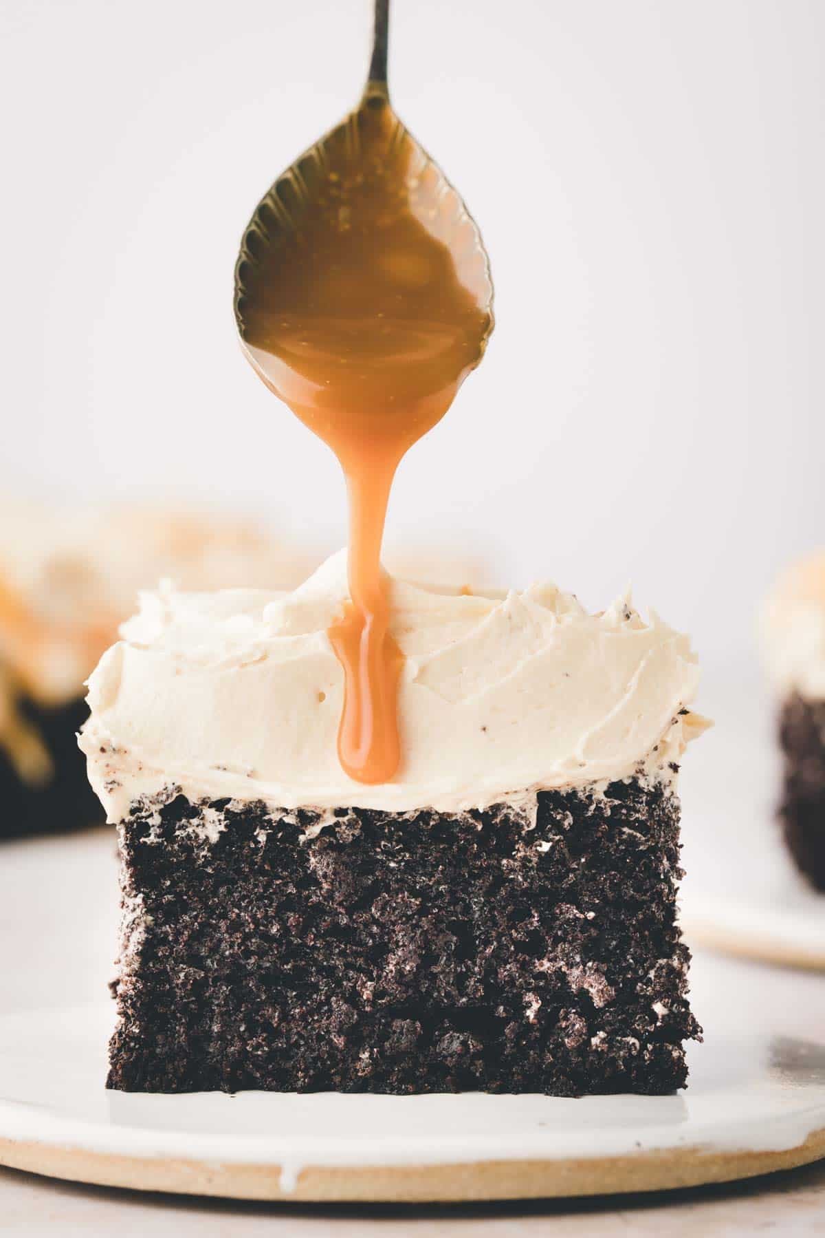 A spoon drizzles caramel sauce over a slice of chocolate cake with light-colored frosting, set on a white plate.
