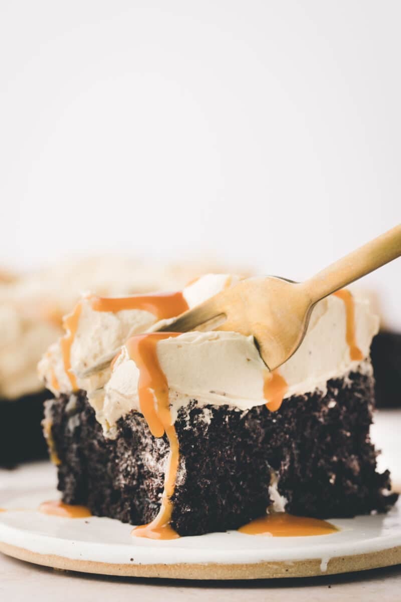 A slice of chocolate cake with caramel drizzle and whipped topping on a plate, with a fork inserted into the cake.