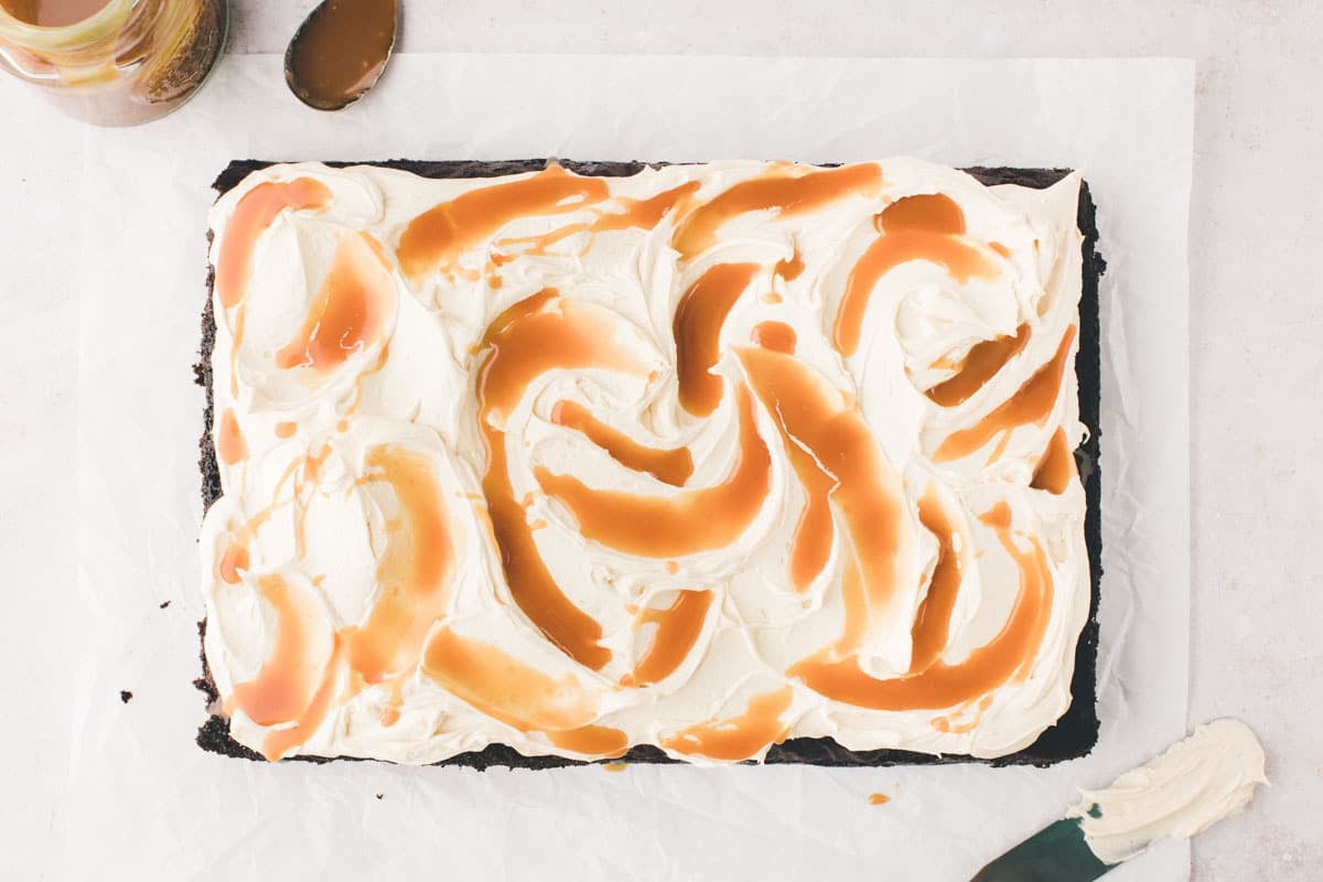 A rectangular chocolate cake topped with swirled white frosting and caramel sauce, placed on parchment paper with a spatula and caramel jar nearby.