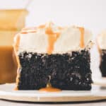 A slice of chocolate cake with white frosting and caramel drizzle sits on a plate, with a jar of caramel sauce in the background.