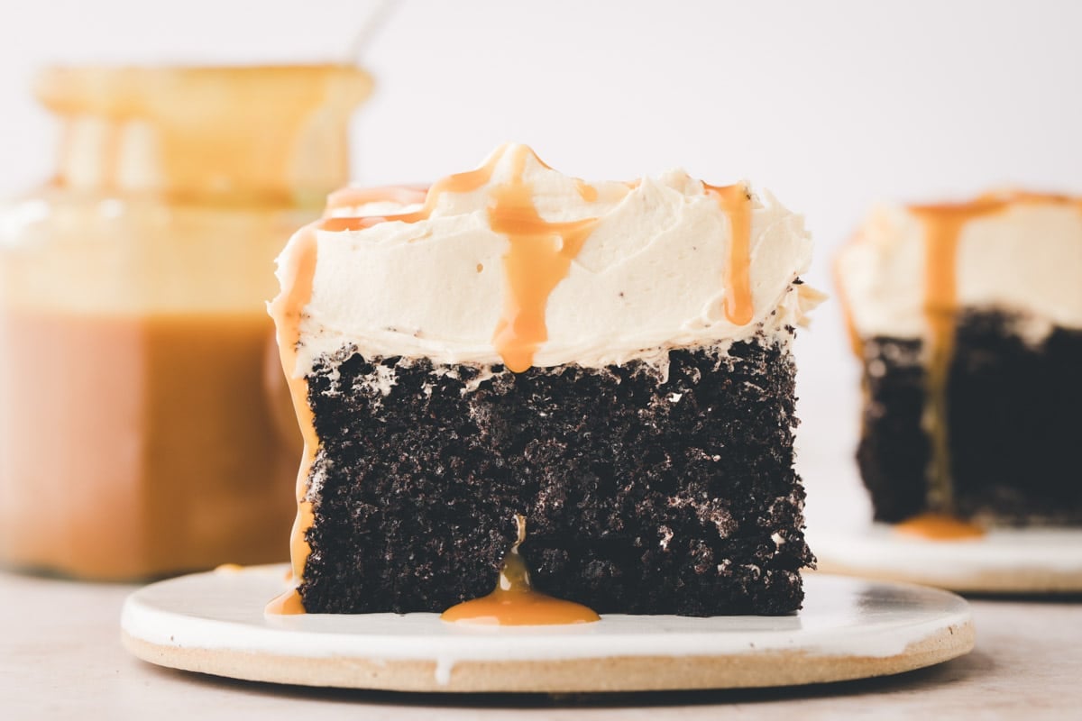 A slice of chocolate cake with white frosting and caramel drizzle sits on a plate, with a jar of caramel sauce in the background.
