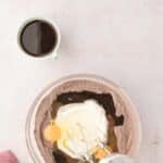 A glass bowl with chocolate cake mix, eggs, oil, and sour cream, next to a cup of coffee; an electric mixer is partially visible in the bowl.