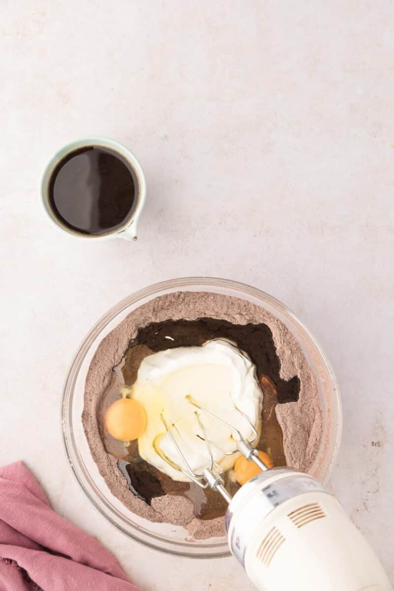 A glass bowl with chocolate cake mix, eggs, oil, and sour cream, next to a cup of coffee; an electric mixer is partially visible in the bowl.