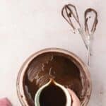 A hand pours liquid from a small cup into a bowl of chocolate batter; two mixer beaters with batter rest nearby on a light surface.