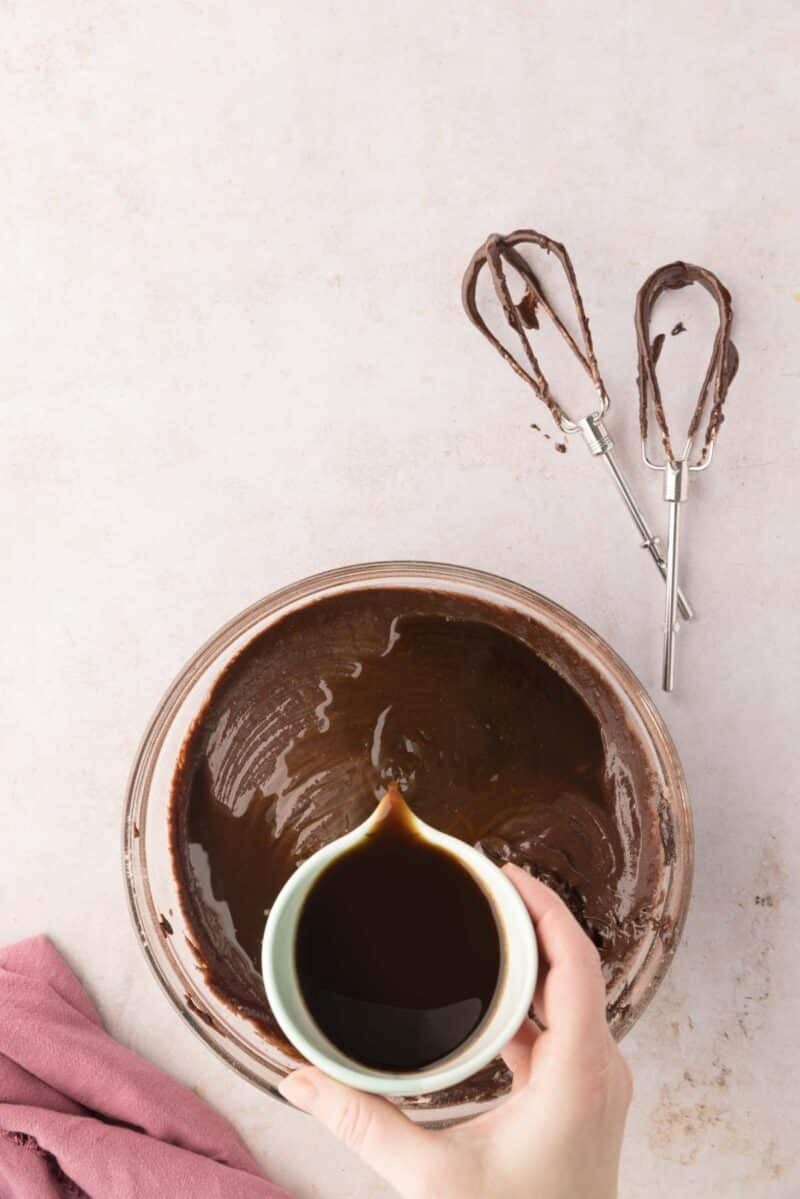 A hand pours liquid from a small cup into a bowl of chocolate batter; two mixer beaters with batter rest nearby on a light surface.