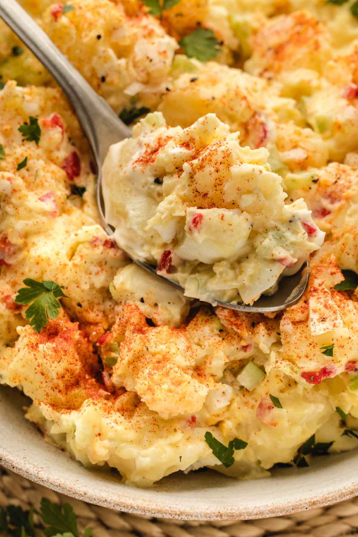 A close-up of a spoonful of potato salad topped with paprika, showing chunks of potatoes, diced vegetables, and parsley in a creamy dressing.