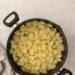 A pot filled with cubed potatoes submerged in water, placed on a light countertop next to a folded gray cloth.