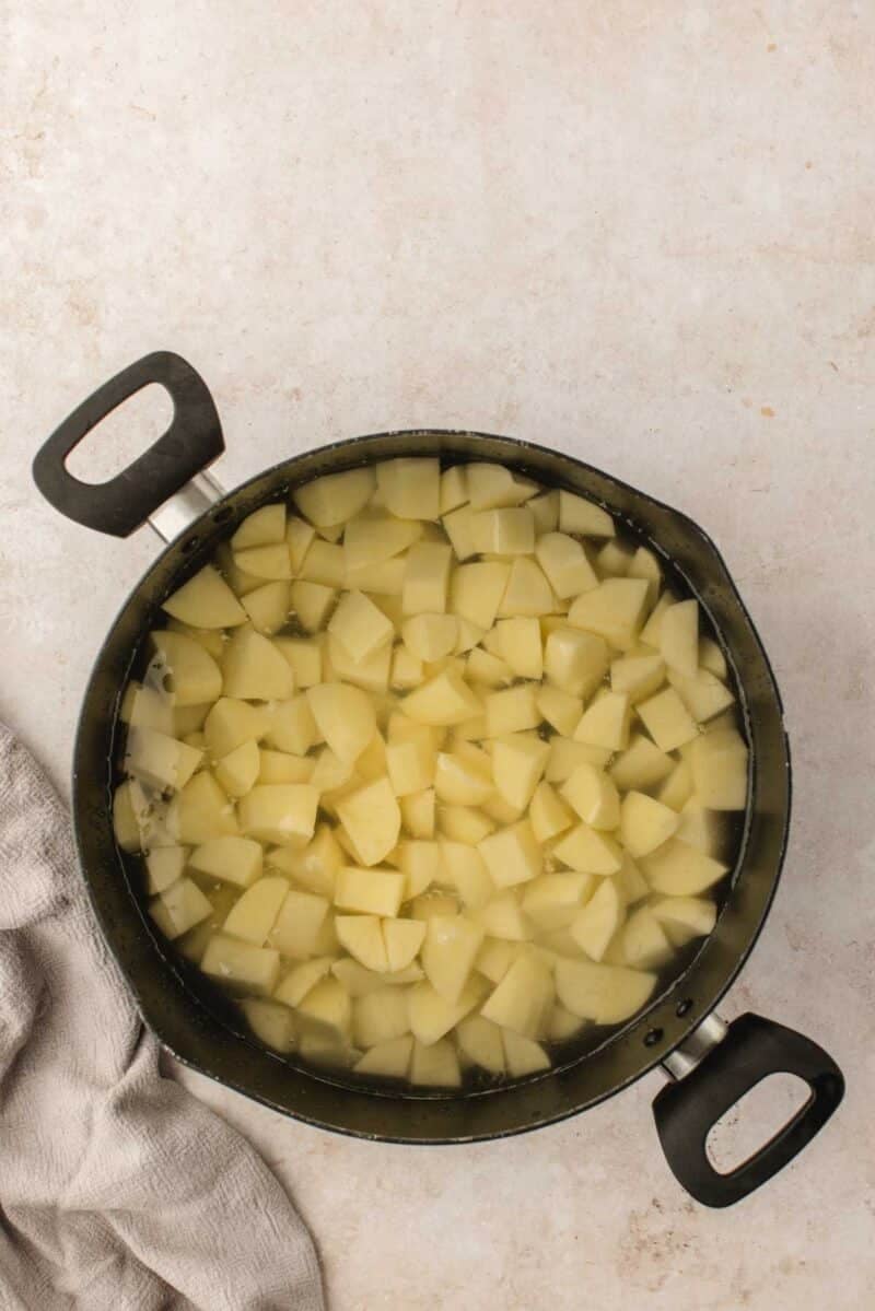 A pot filled with cubed potatoes submerged in water, placed on a light countertop next to a folded gray cloth.