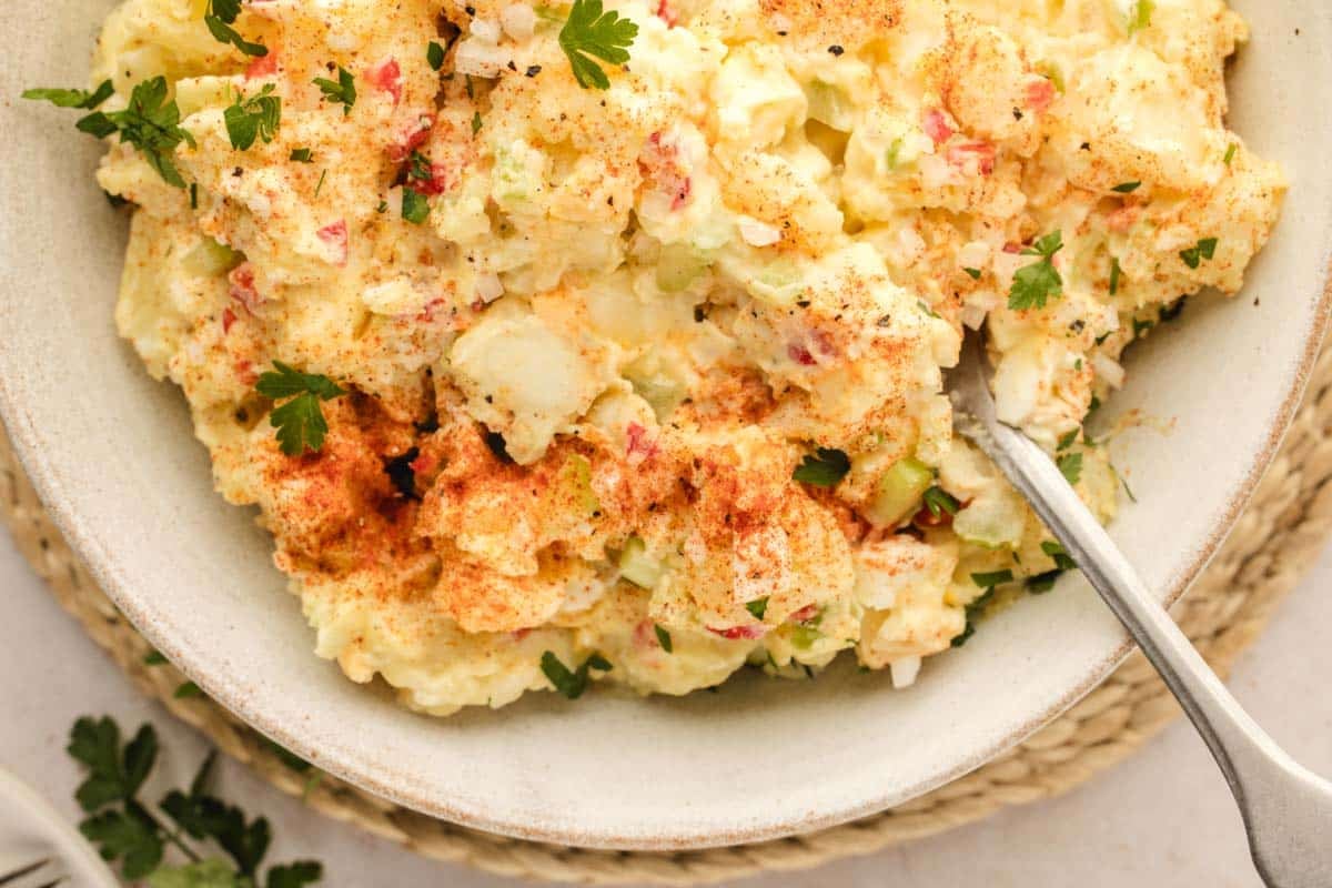 A bowl of potato salad garnished with chopped parsley and paprika, with a spoon resting inside.