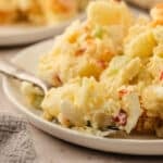 A close-up of a plate of potato salad with visible chunks of potatoes, eggs, and vegetables, with a fork resting on the plate.