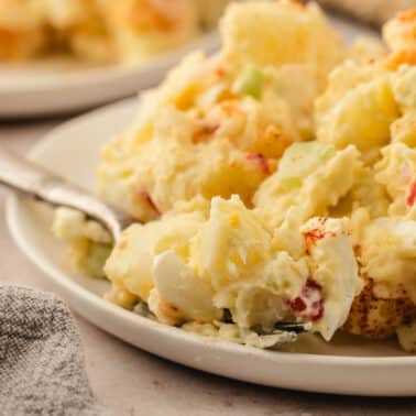 A close-up of a plate of potato salad with visible chunks of potatoes, eggs, and vegetables, with a fork resting on the plate.