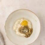 A white bowl contains mayonnaise, Dijon mustard, grated Parmesan cheese, pesto, and ground black pepper on a light surface.