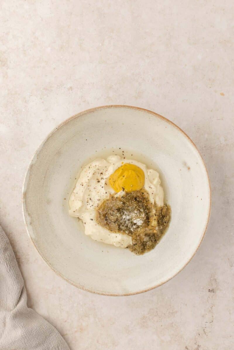 A white bowl contains mayonnaise, Dijon mustard, grated Parmesan cheese, pesto, and ground black pepper on a light surface.