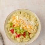 Bowl of potato salad topped with chopped hard-boiled eggs, diced celery, shredded cheese, and bits of red pepper on a light surface.