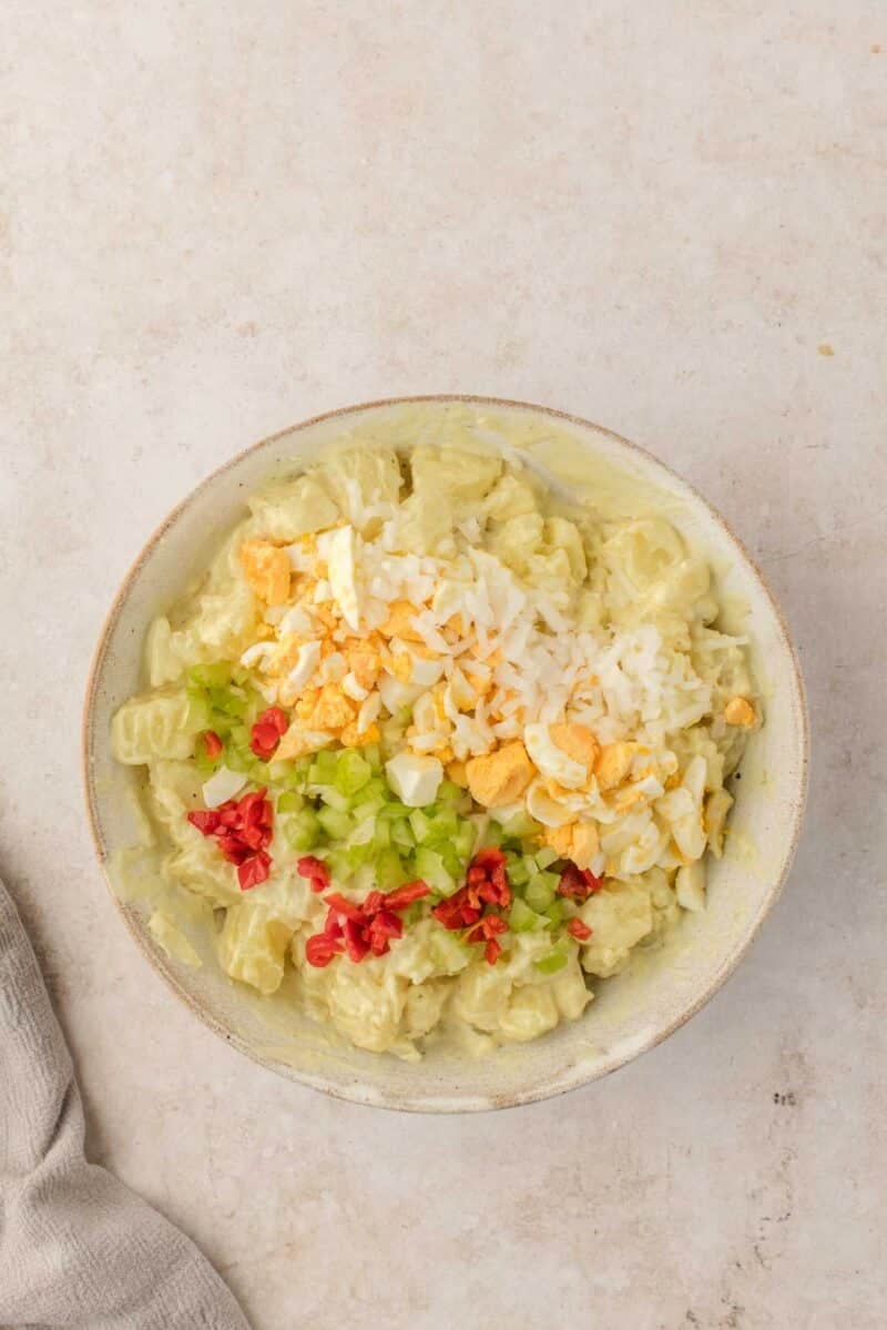 Bowl of potato salad topped with chopped hard-boiled eggs, diced celery, shredded cheese, and bits of red pepper on a light surface.
