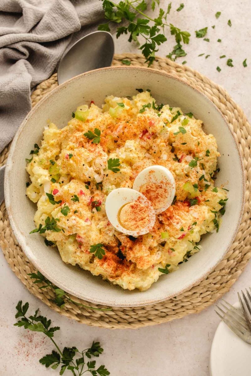 A bowl of potato salad garnished with sliced hard-boiled egg, paprika, and chopped parsley, placed on a woven mat with utensils and a napkin nearby.