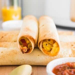 Four breakfast taquitos filled with scrambled eggs, cheese, and bacon are arranged on a board; a small bowl of salsa and a glass of orange juice are visible nearby.