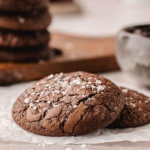 Two chocolate brownie cookies topped with coarse sea salt rest on white parchment paper, with a blurred stack of cookies and a bowl in the background.