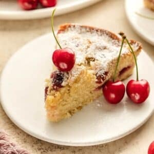 A slice of cherry almond cake dusted with powdered sugar is served on a white plate, garnished with three fresh cherries.