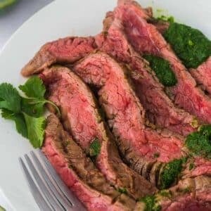 Sliced medium-rare steak served on a white plate with green sauce drizzled on top and a sprig of fresh cilantro, next to a fork.