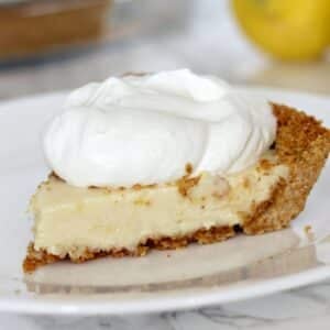 A slice of old-fashioned lemon icebox pie with a graham cracker crust, creamy filling, and a generous dollop of whipped cream on top, served on a white plate.