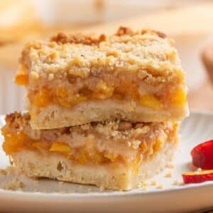 Two peach crumb bars are stacked on a white plate, showing layers of fruit filling and crumb topping, with a peach slice nearby.